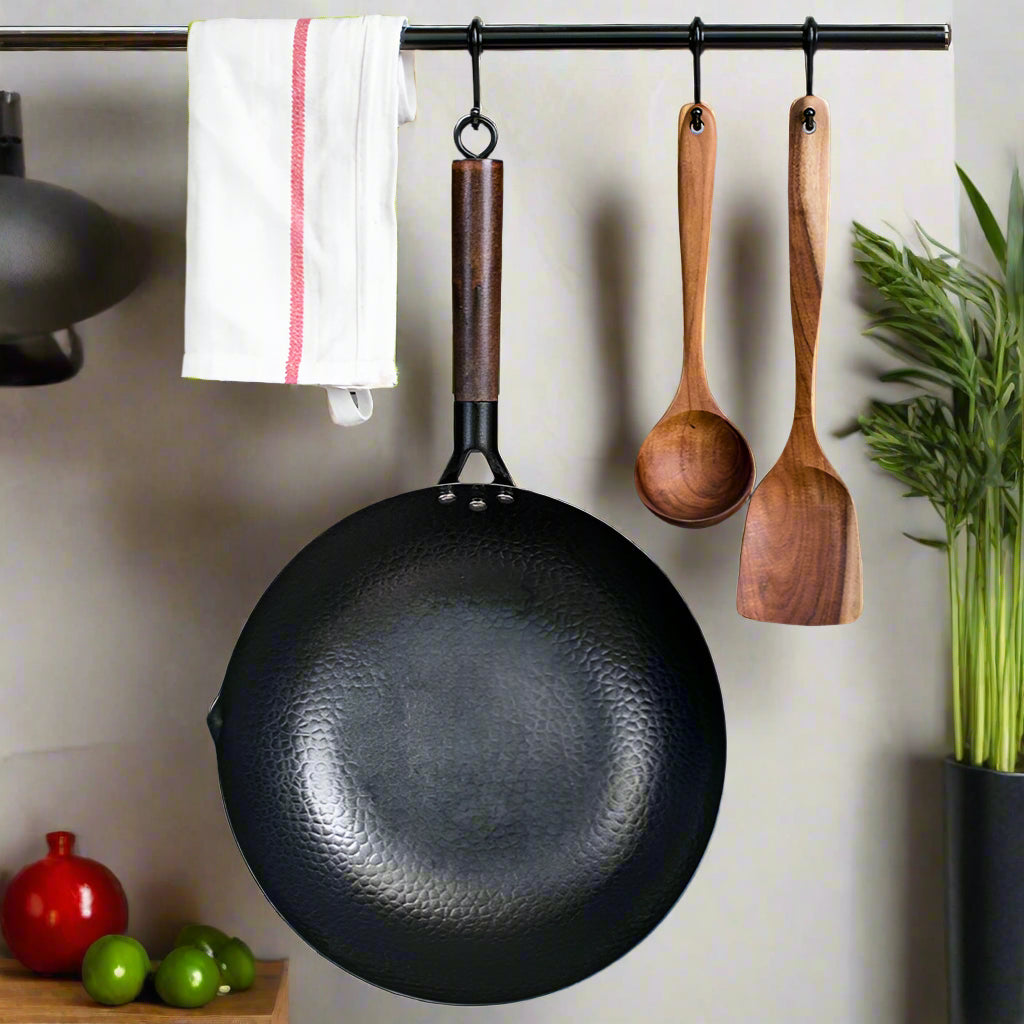 Black wok with wooden handle and two wooden spoons hanging on a metal rod against a gray tiled wall.
