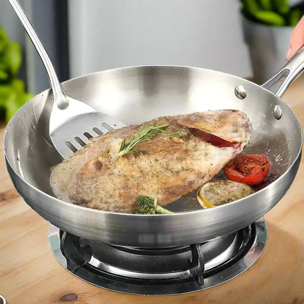 Stainless steel frying pan with cooked fish and vegetables on a stove, brand 'KEMORELA' visible.