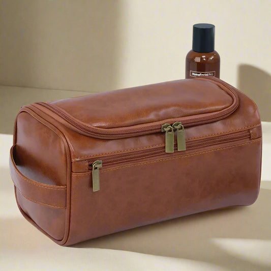Brown leather toiletry bag with gold zippers on a white background, next to a bottle and razor.