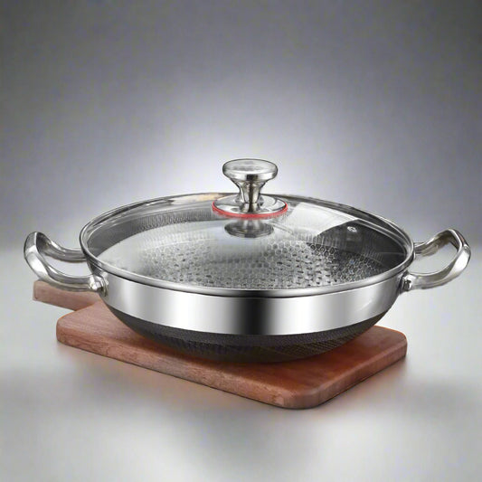 Stainless steel wok with glass lid on a kitchen counter with food and utensils.